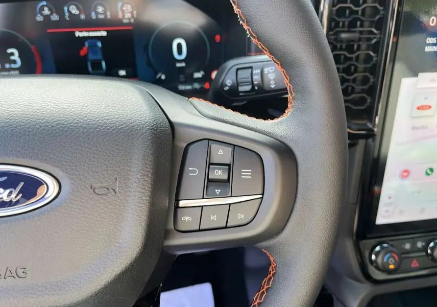 Gros plan sur le volant cuir noir avec surpiqûres orange du Ford Ranger Wildtrak 2026, tableau de bord numérique et écran tactile visibles.