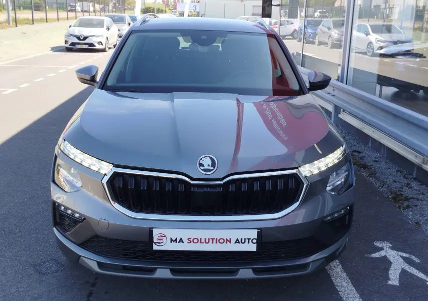 Vue frontale d'un Skoda Kamiq gris graphite métal 2024 avec calandre noire et feux LED allumés, stationné devant un showroom.