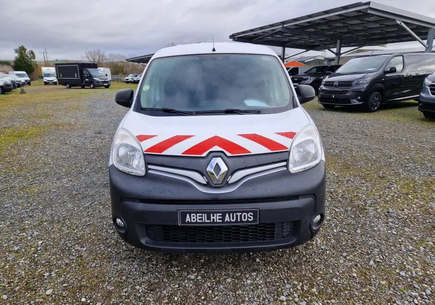 Vue de face d'un Renault Kangoo blanc avec bandes rouges réfléchissantes sur le capot, stationné sur un sol gravillonné.