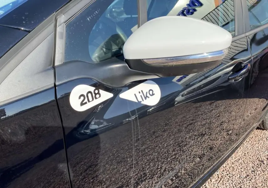 Vue latérale gauche rapprochée d’une Peugeot 208 noire avec rétroviseur blanc et logos 208 Like sur la porte avant.