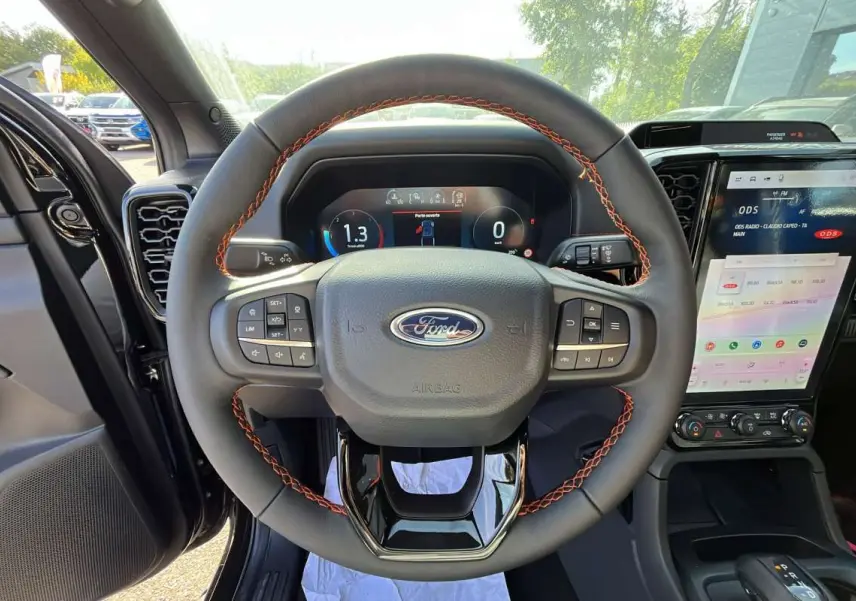 Vue intérieure centrée sur le volant cuir noir avec surpiqûres orange du Ford Ranger Wildtrak 2026, écran tactile visible à droite.