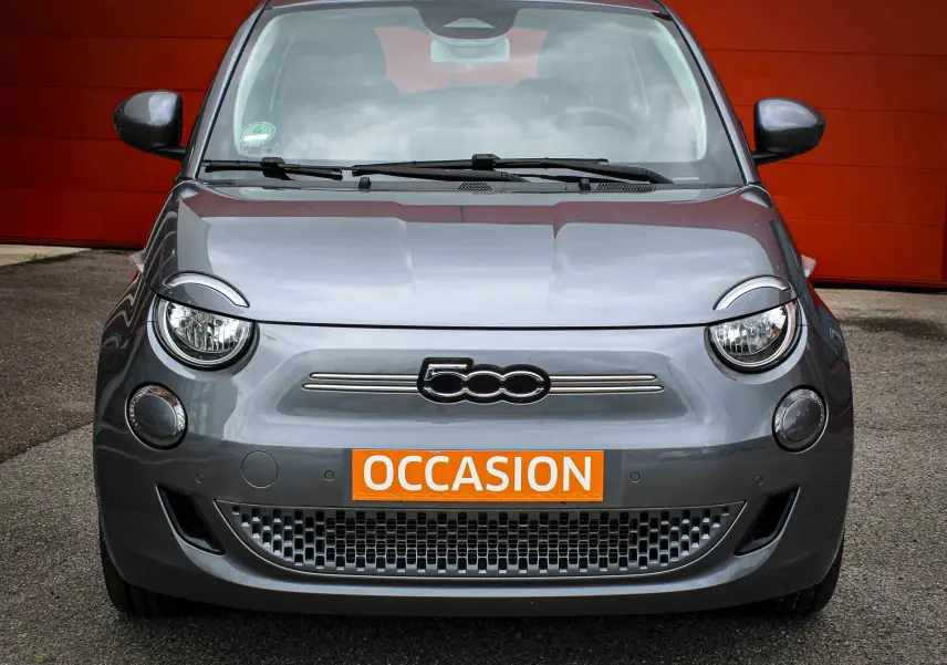 Vue frontale d'une FIAT 500 électrique gris minéral métal avec plaque orange "OCCASION" sur fond rouge.