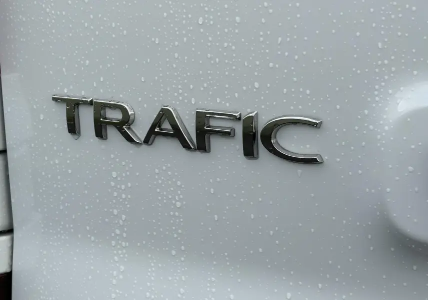 Gros plan sur l’inscription TRAFIC en chrome sur la porte arrière d’un Renault Trafic blanc avec gouttes d’eau.