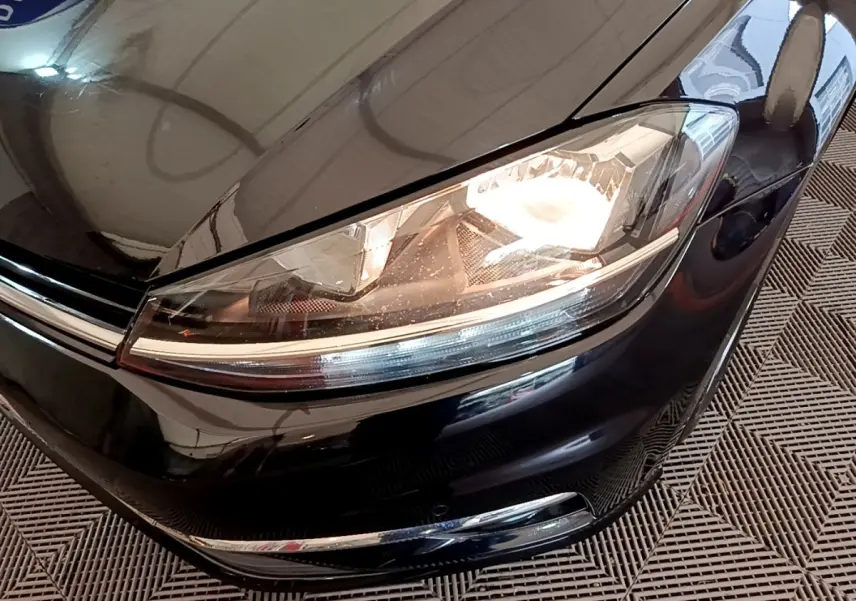 Gros plan sur le phare avant droit allumé d’une Volkswagen Golf noire, avec calandre chromée visible.