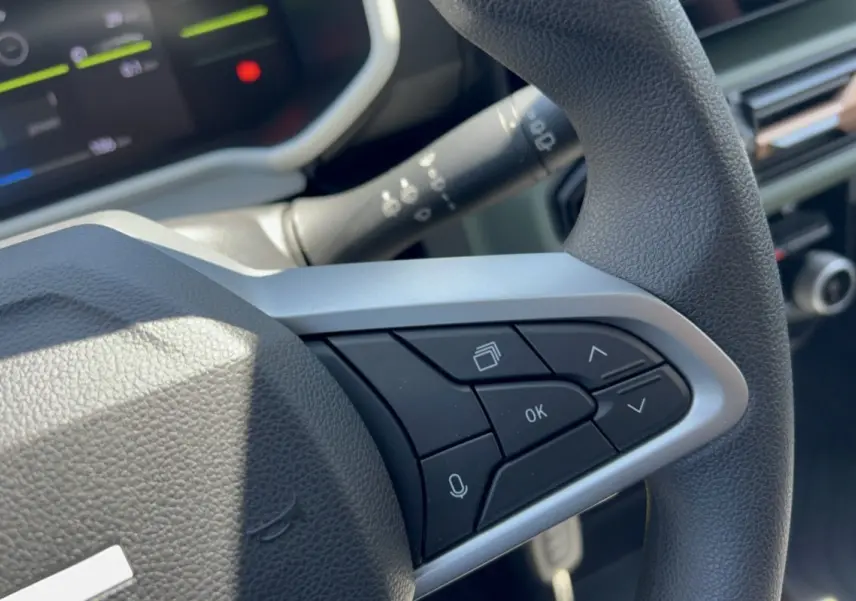 Gros plan sur les commandes au volant côté droit du Dacia Spring gris éclair, avec tableau de bord numérique en arrière-plan.