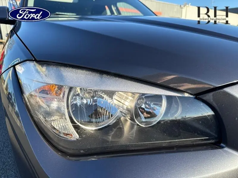 Gros plan sur le phare avant gauche du BMW X1 Mineralgrau 2013, mettant en valeur la teinte minérale et les détails du bloc optique.