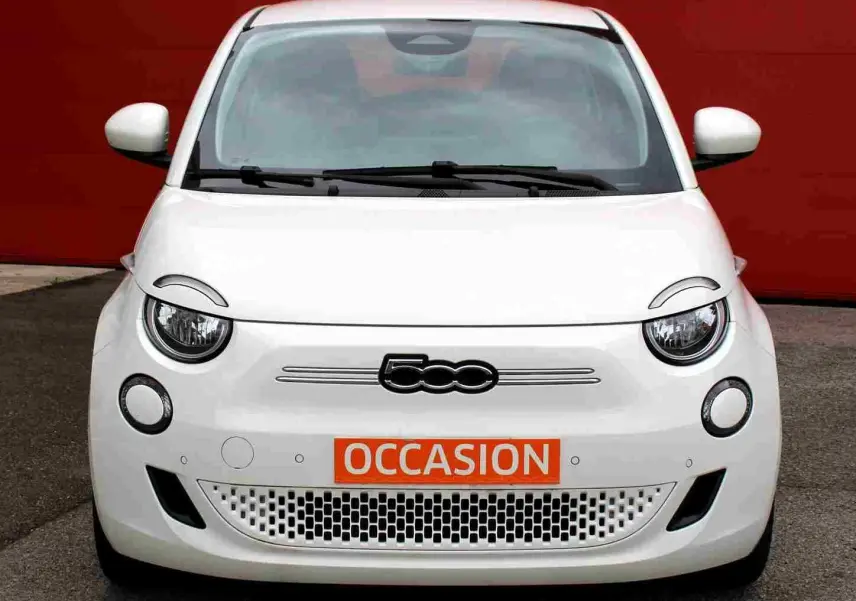 Vue avant d'une FIAT 500 électrique blanche avec logo distinctif et plaque orange "OCCASION" devant un mur rouge.