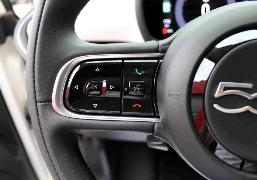 Gros plan sur les commandes à gauche du volant noir de la Fiat 500 électrique blanche, avec logo 500 visible.