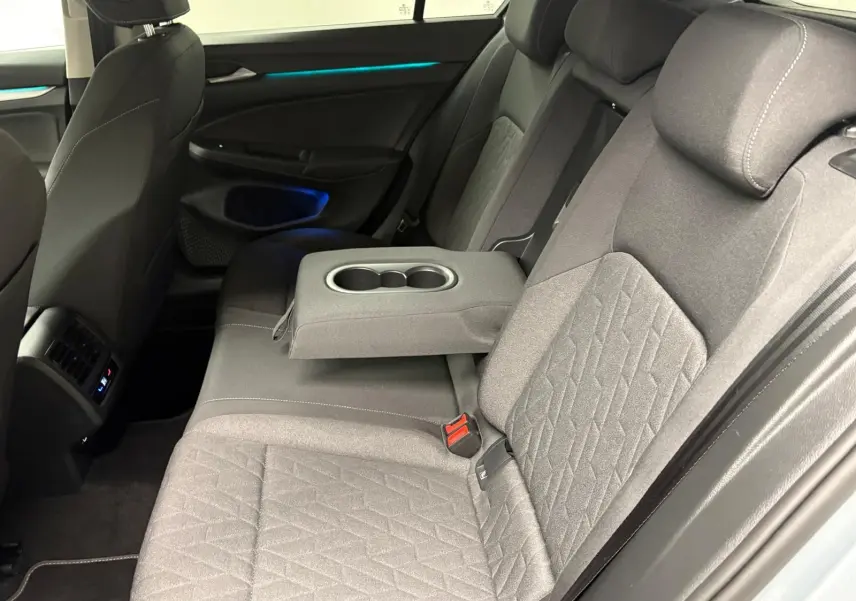 Vue intérieure arrière droite de la Volkswagen Golf 1.5 TSI 2025, sièges gris avec accoudoir central et éclairage d'ambiance bleu.