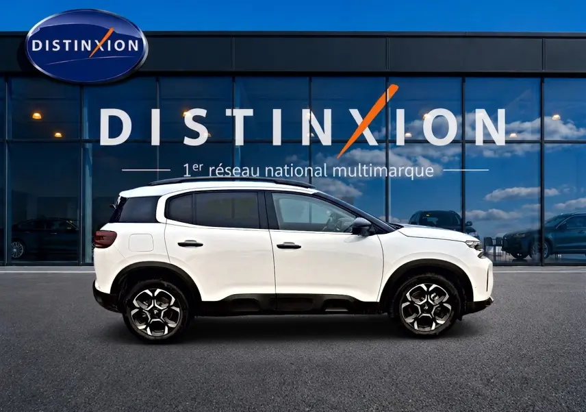 Profil droit du Citroën C5 Aircross blanc 2025 avec jantes alu et barres de toit visibles devant une vitrine.