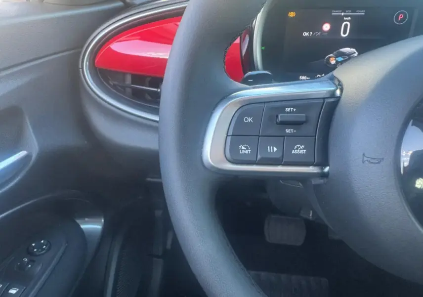Vue intérieure rapprochée du volant et tableau de bord d’une FIAT 600 rouge, avec commandes au volant visibles côté gauche.