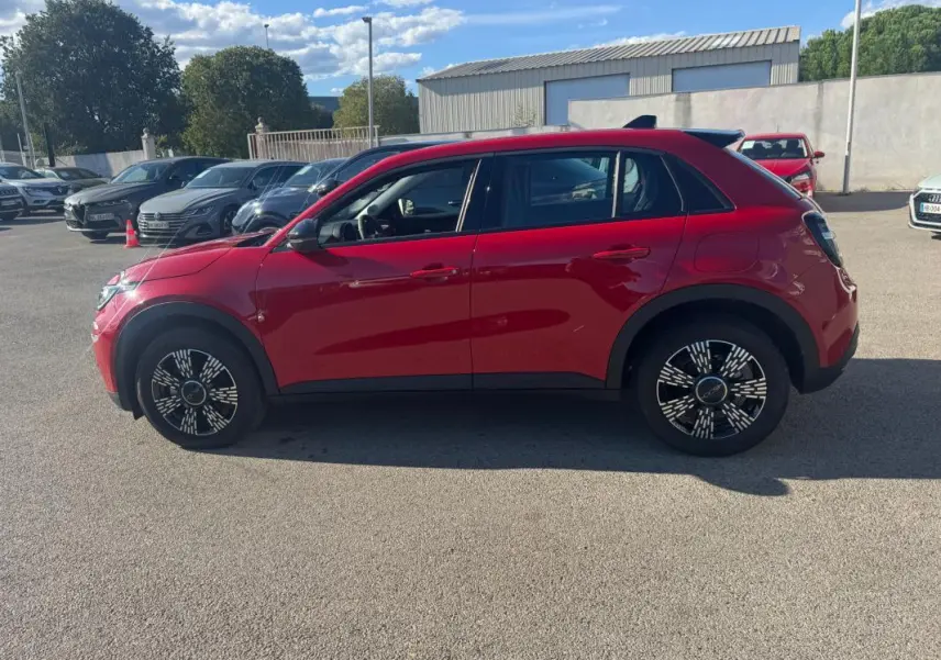 Vue de profil côté gauche d'une FIAT 600 T-Gen 3 rouge 2025 avec jantes au design moderne sur un parking extérieur.