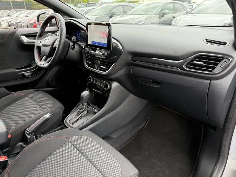 Vue intérieure côté passager du Ford Puma 2024 ST-Line, tableau de bord noir avec écran tactile et sièges tissu gris à surpiqûres rouges.