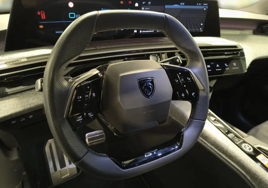 Vue rapprochée du volant compact noir perforé avec badge Peugeot et inscription GT, tableau de bord numérique en arrière-plan.