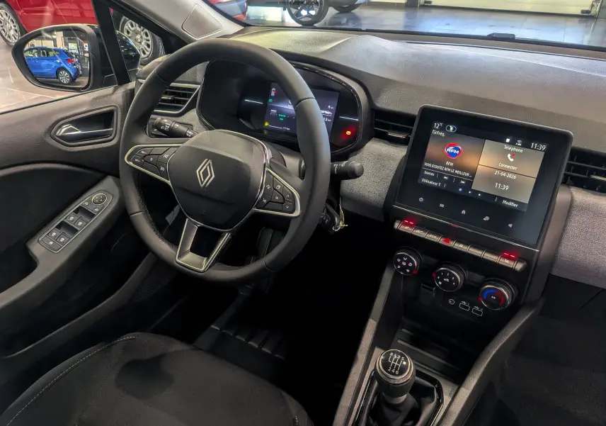 Vue intérieure côté conducteur de la Renault Clio V 2025, tableau de bord moderne avec écran tactile et levier de vitesse manuel.