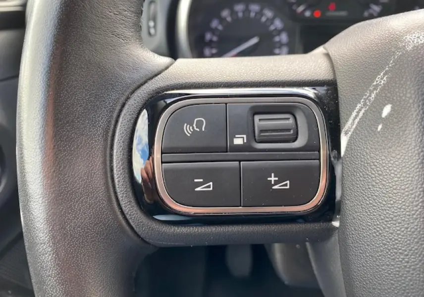 Gros plan sur les commandes de volume et téléphone au volant d'une Citroën C3 gris clair, intérieur visible en arrière-plan flou.