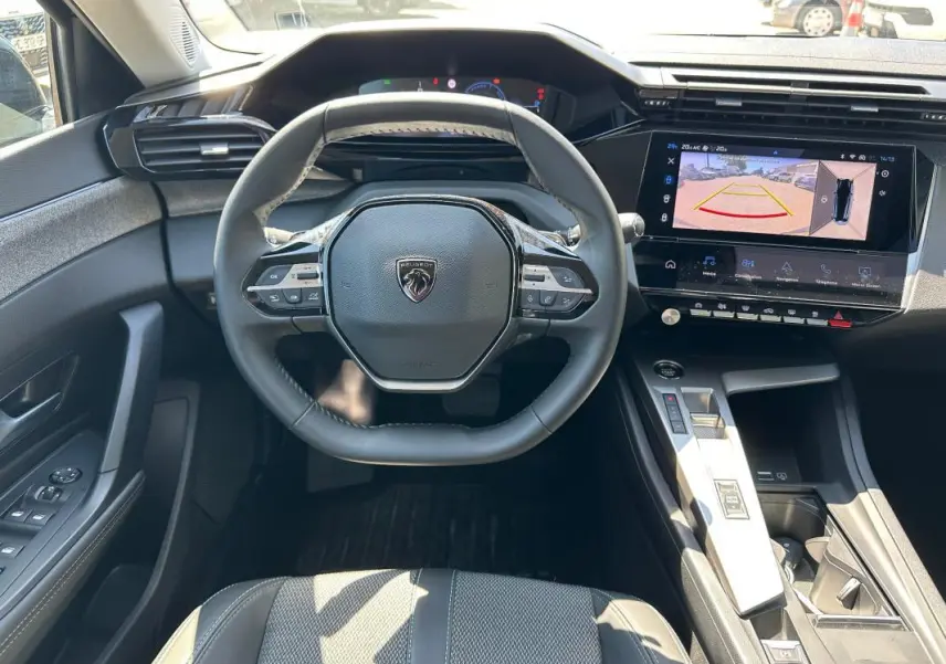 Vue intérieure centrée sur le volant et la console d'une Peugeot 308 SW Hybrid noire avec écran tactile et caméra de recul affichée.