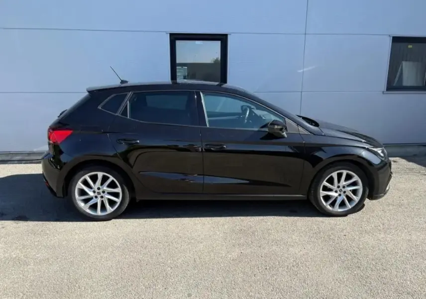 Profil droit d’une SEAT Ibiza 1.0 TSI 95 FR noire, avec jantes alliage argentées, stationnée devant un mur gris.