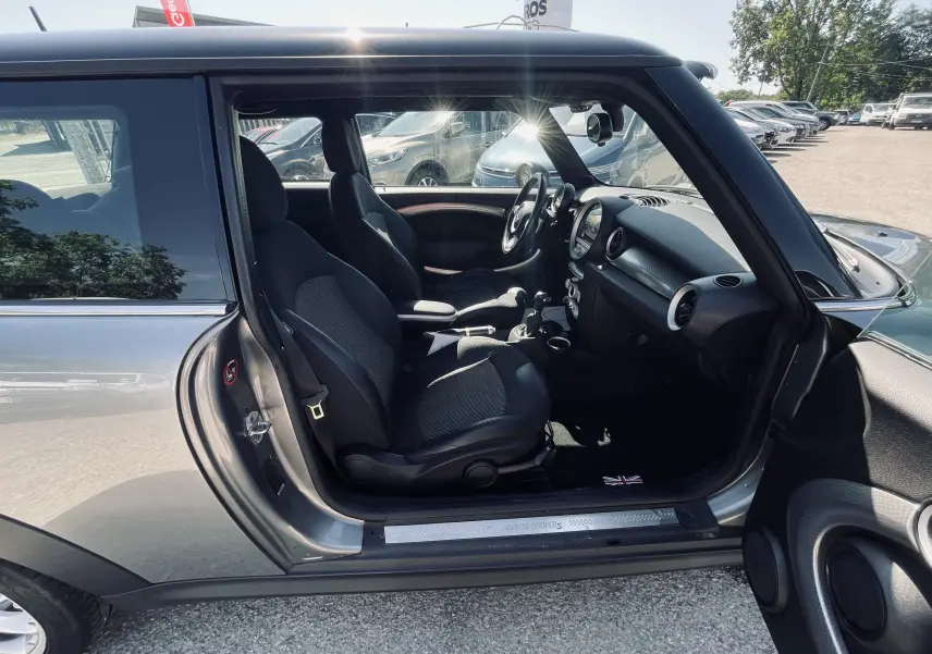 Intérieur côté gauche d'une MINI Cooper S gris 2008, avec sièges noirs et tableau de bord visible au soleil.
