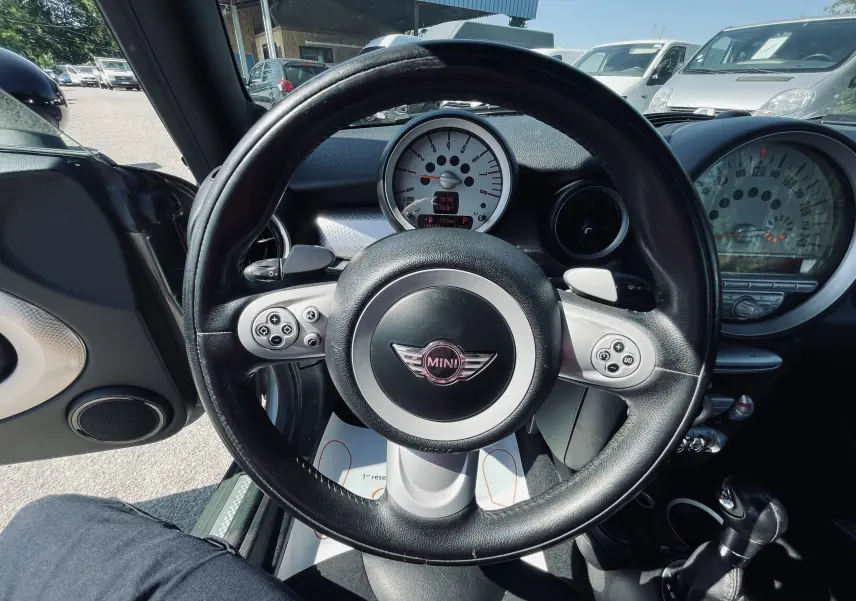 Vue intérieure centrée sur le volant noir d'une MINI Cooper S 2008 avec tableau de bord et levier de boîte auto visibles.