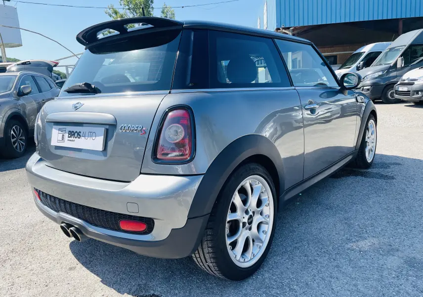 Vue 3/4 arrière droite d'une MINI Cooper S grise avec becquet noir et jantes argentées sur parking extérieur.