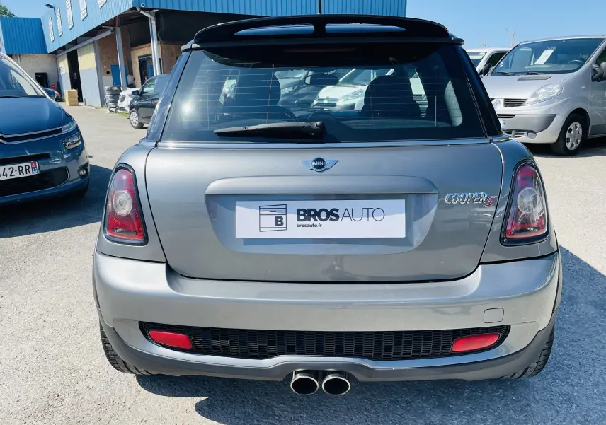 Vue arrière d'une MINI Cooper S grise 2008 avec double sortie d'échappement et becquet noir sur parking.
