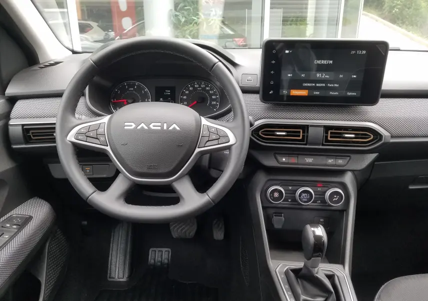 Vue intérieure centrée sur le volant DACIA et le tableau de bord avec écran tactile et commandes de climatisation.