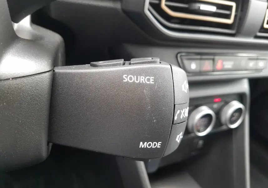 Commande au volant noir avec boutons source et mode, tableau de bord et commandes de climatisation en arrière-plan dans une Dacia Sandero Stepway