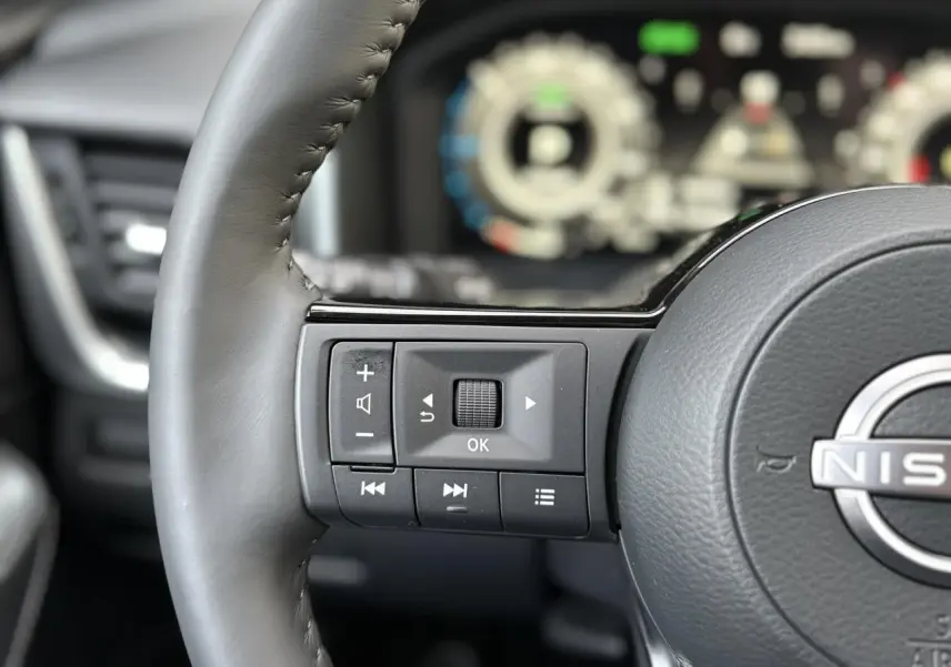 Gros plan sur les commandes gauche du volant cuir gris du Nissan Qashqai III e-Power avec tableau de bord numérique flou en arrière-plan.