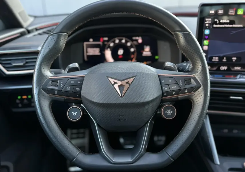 Vue rapprochée du volant cuir perforé noir avec logo CUPRA cuivré et commandes intégrées, tableau de bord digital en arrière-plan.