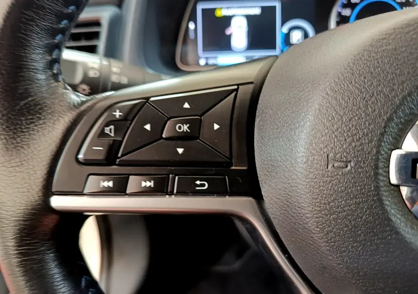 Gros plan sur les commandes gauche du volant cuir noir du Nissan Leaf blanc, avec affichage numérique flou en arrière-plan.