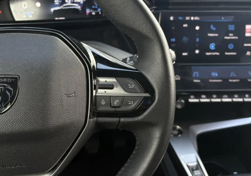 Gros plan sur le volant en cuir noir de la Peugeot 308 2024 avec commandes intégrées et tableau de bord numérique en arrière-plan.