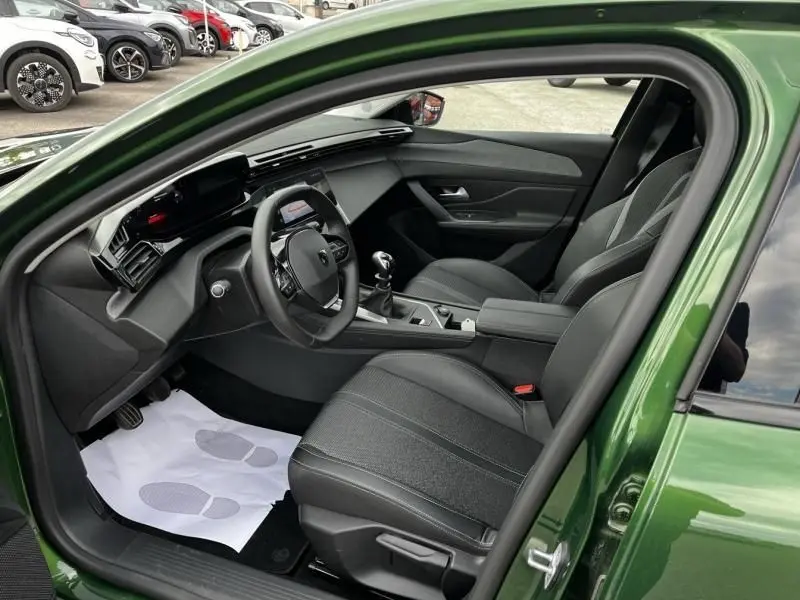 Vue intérieure côté conducteur de la Peugeot 308 1.2 PureTech 130 Allure 2023 en vert olivine, avec volant et boîte manuelle visibles.