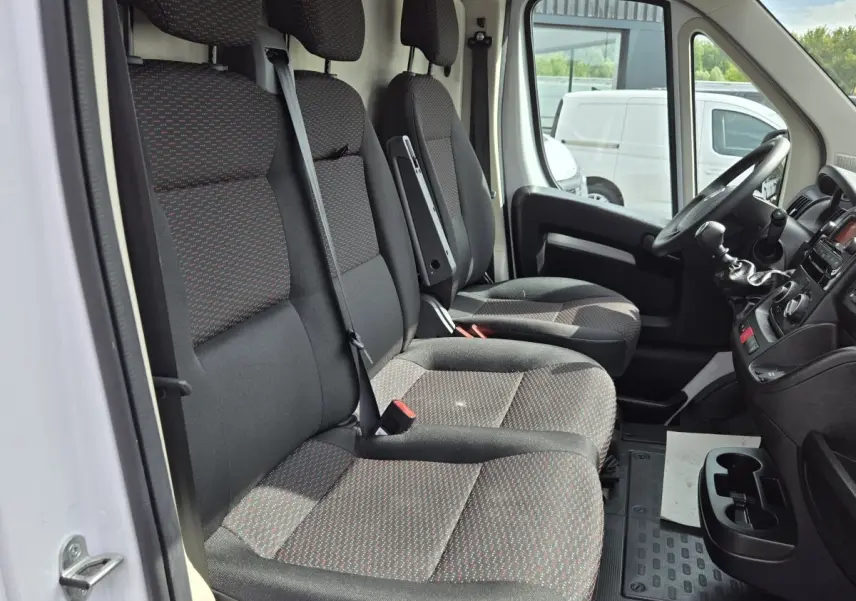 Vue intérieure côté conducteur du Fiat Ducato Fourgon blanc 2023, montrant les sièges avant en tissu noir à motifs rouges.