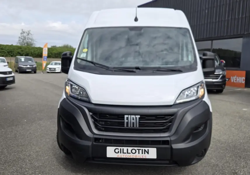 Vue frontale d'un FIAT Ducato Fourgon blanc avec calandre noire et phares allumés, sur parking extérieur.