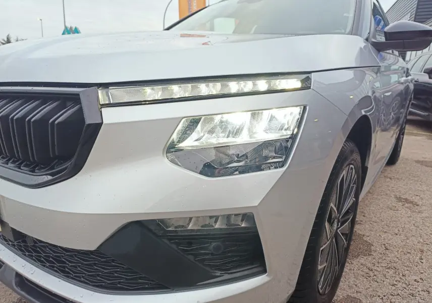 Vue 3/4 avant droit d'un Skoda Kamiq 2025 argent avec toit noir et phares LED allumés.