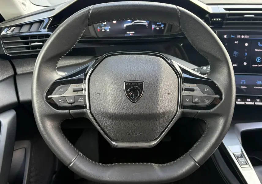 Vue rapprochée du volant cuir noir au design hexagonal de la Peugeot 308 2024 avec tableau de bord numérique en arrière-plan.
