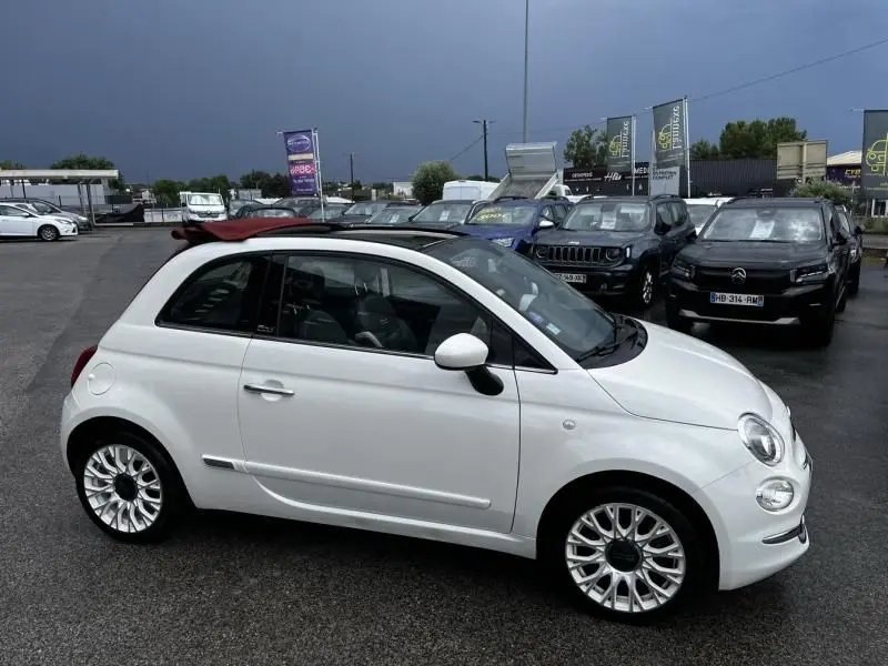 Vue de profil côté gauche d'une FIAT 500C blanche avec toit ouvrant partiellement ouvert sur un parking.