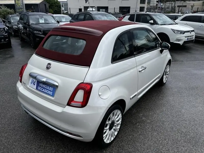 Vue 3/4 arrière droite d'une Fiat 500C blanche avec toit ouvrant en toile bordeaux, stationnée sur parking humide.