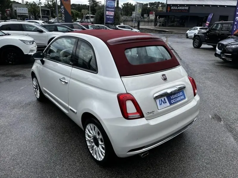 Vue 3/4 arrière droite d'une Fiat 500C blanche avec toit ouvrant rouge bordeaux sur parking humide.