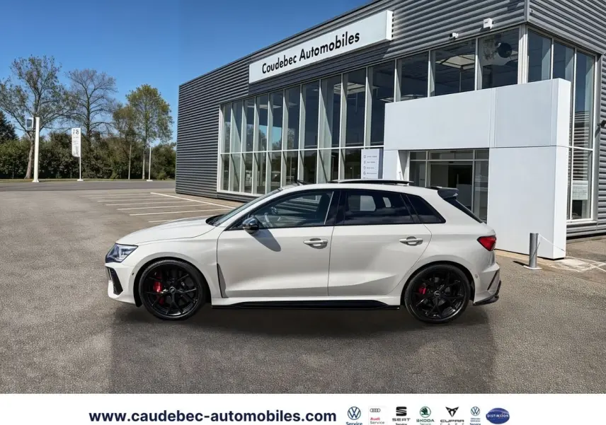 Audi RS3 Sportback gris vue de profil droit, jantes noires et étriers de frein rouges, devant un bâtiment moderne.