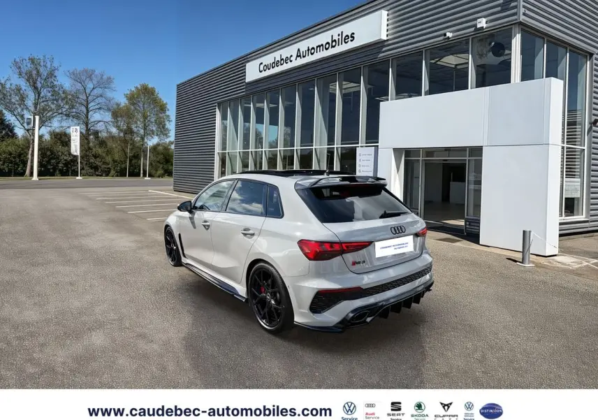 Audi RS3 Sportback gris vue 3/4 arrière droit, jantes noires, toit panoramique et becquet carbone visible.