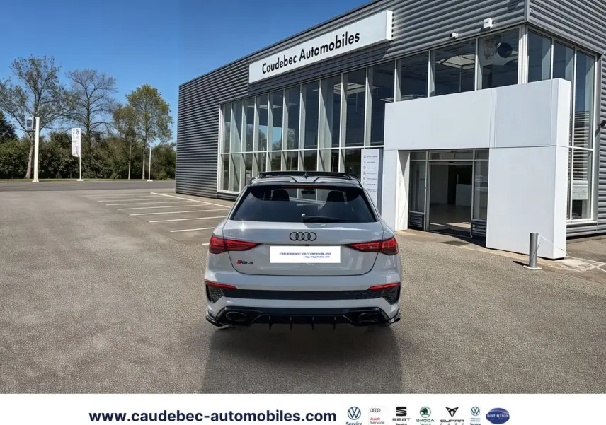 Vue arrière d'une Audi RS3 Sportback grise avec diffuseur noir et feux arrière LED, stationnée devant un bâtiment moderne.
