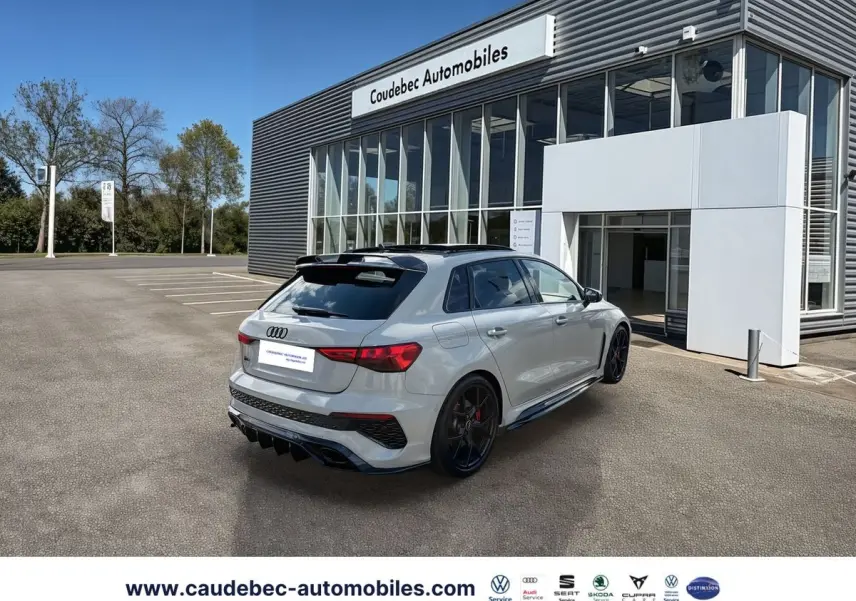 Audi RS3 Sportback grise vue 3/4 arrière droit, jantes noires mat et détails carbone visibles, stationnée devant un showroom.
