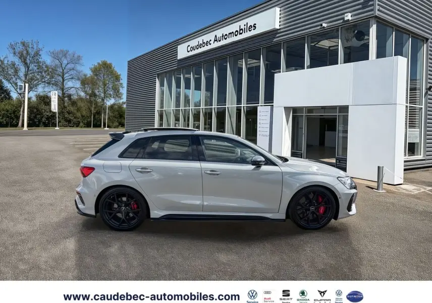 Audi RS3 Sportback grise vue de profil côté gauche, avec jantes noires et étriers de frein rouges, devant un showroom.