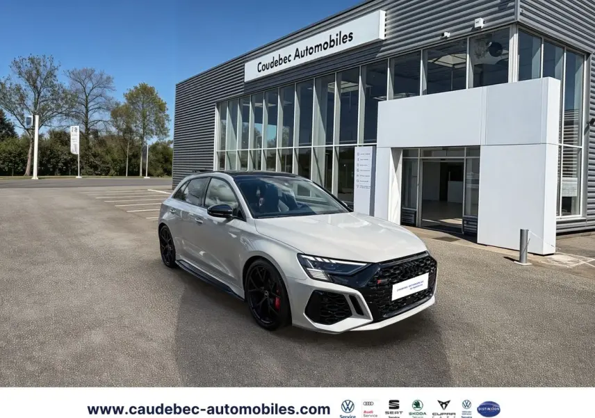 Audi RS3 Sportback gris vue 3/4 avant droit, jantes noires et étriers de frein rouges devant un showroom moderne.