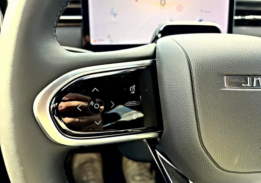 Gros plan sur les commandes multifonctions à gauche du volant gris du JAECOO J7, avec écran tactile en arrière-plan.