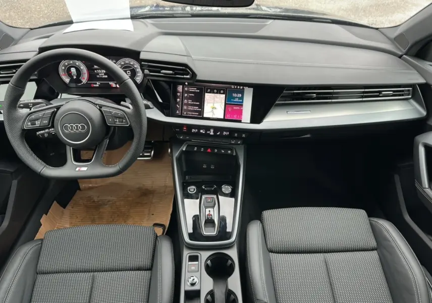 Intérieur Audi A3 2025 vu de face, volant cuir méplat, écran tactile central et sièges sport tissu cuir gris.