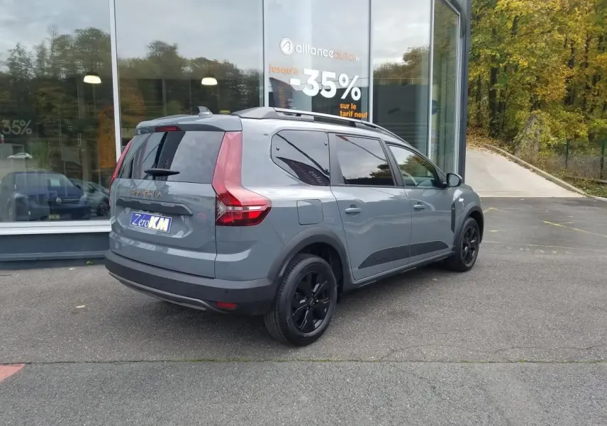 Vue 3/4 arrière droite d'un Dacia Jogger gris urbain avec barres de toit et vitres teintées, stationné devant une vitrine.