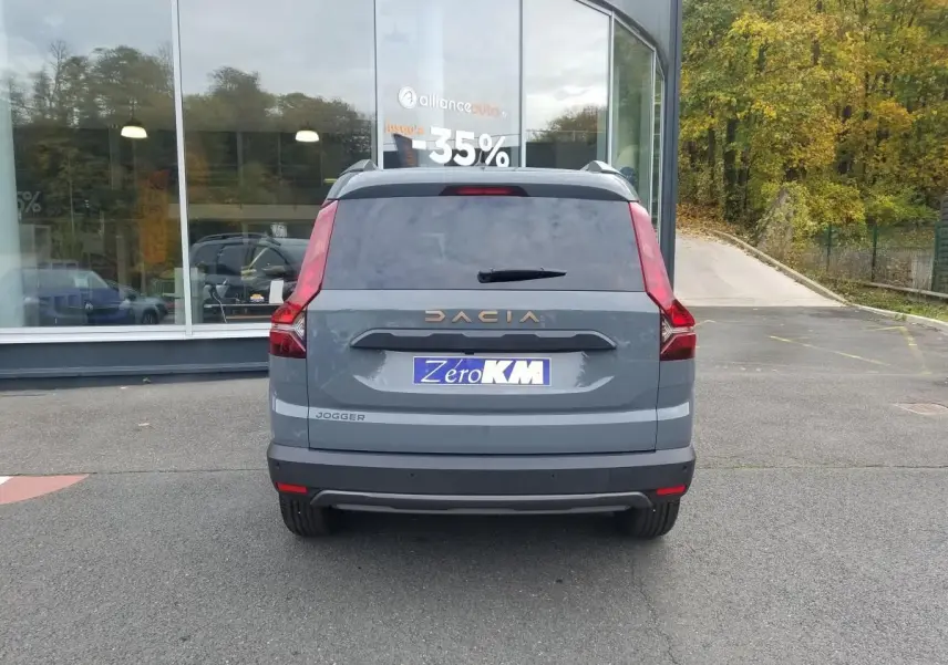 Vue arrière d'un Dacia Jogger gris urbain 2025 avec feux verticaux et logo doré sur un parking devant une vitrine.